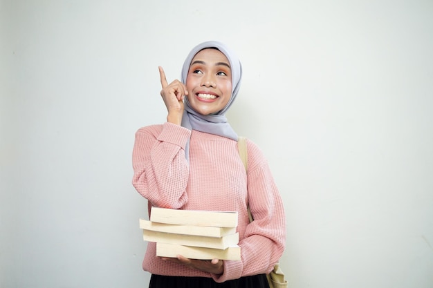 Podekscytowana młoda azjatycka muzułmańska studentka w różowym swetrze z torbą i trzymającą książkę