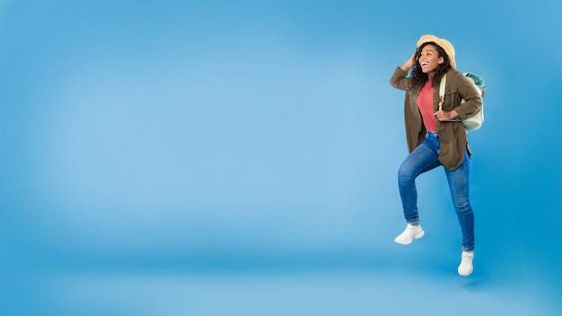 Podekscytowana młoda czarna kobieta z plecakiem biegnącym szczęśliwie nad jej letnimi wakacjami w niebieskim studio
