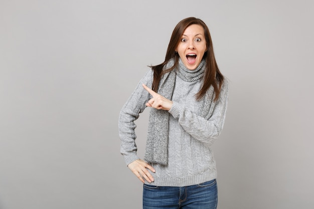 Podekscytowana młoda kobieta w szary sweter szalik wskazując palcem na bok utrzymując otwarte usta na białym tle na szarym tle. Zdrowa moda styl życia ludzie emocje koncepcja zimnej pory roku. Makieta miejsca na kopię.