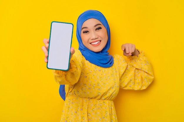 Podekscytowana młoda piękna azjatycka muzułmanka pokazująca pusty ekran telefonu komórkowego na białym tle na żółtym tle