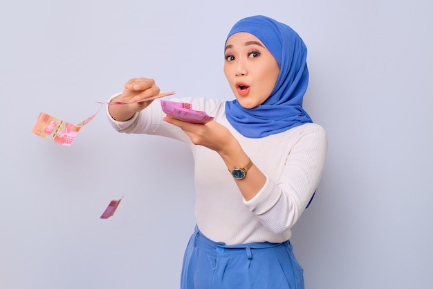 Podekscytowana młoda piękna azjatycka muzułmanka rzucająca banknotami pieniędzy na białym tle na białym tle