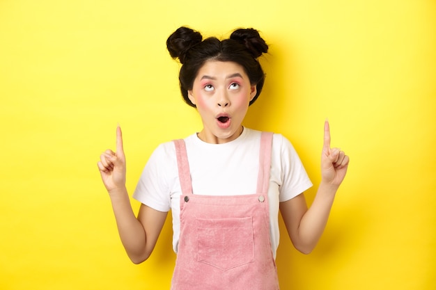 Podekscytowana nastoletnia studentka z Azji z efektownym makijażem, dysząca zdumiona, wskazująca palcami w górę, pokazująca reklamę, żółta.