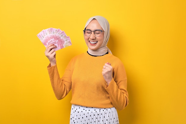 Podekscytowana piękna Azjatka ubrana w hidżab trzymająca banknoty i świętująca sukces odizolowana na żółtym tle