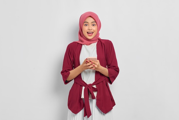Podekscytowana piękna Azjatka w luźnej koszuli i hidżabie, korzystająca z telefonu komórkowego, otrzymała dobre wieści odizolowane na białym tle. Koncepcja życia religijnego ludzi