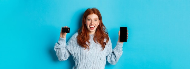 Podekscytowana ruda dziewczyna pokazująca ekran telefonu komórkowego i kartę kredytową pokazującą sklep internetowy lub aplikację