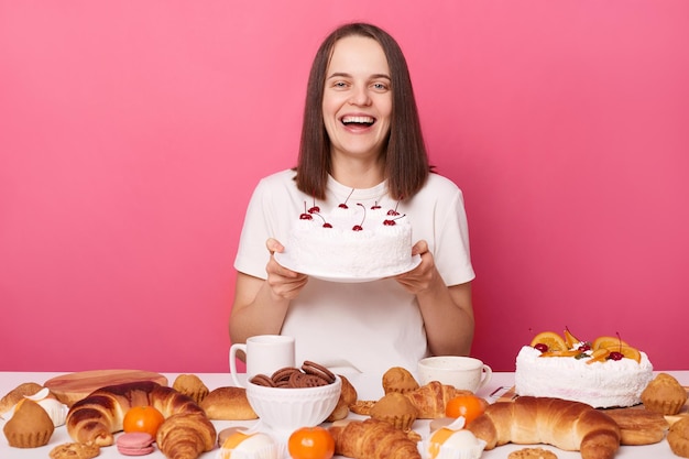 Podekscytowana wesoła kobieta ubrana w białą koszulkę siedząca przy świątecznym stole z różnymi deserami wyizolowanymi na różowym tle pozuje z ciastem w rękach po oszukiwanym posiłku