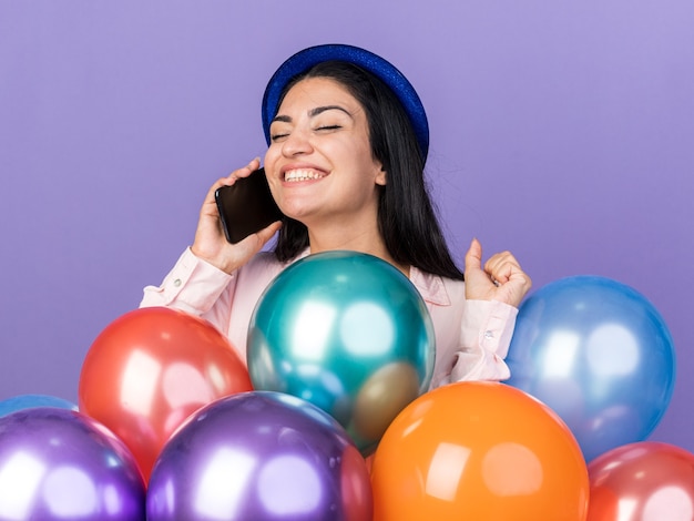 Podekscytowana z zamkniętymi oczami młoda piękna dziewczyna w kapeluszu imprezowym stojąca za balonami rozmawia przez telefon pokazując gest tak na niebieskiej ścianie