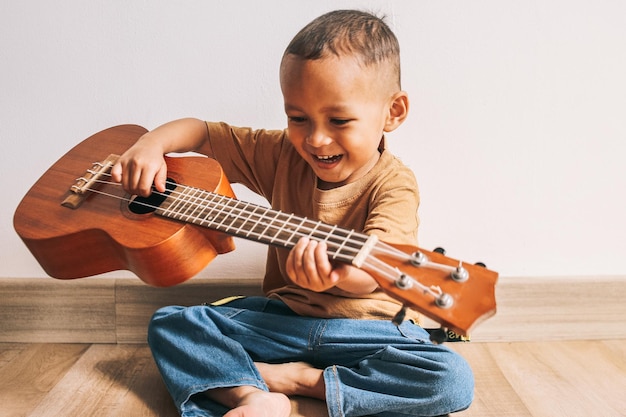 Podekscytowane azjatyckie małe dziecko siedzi na podłodze i gra na gitarze ukulele w domu