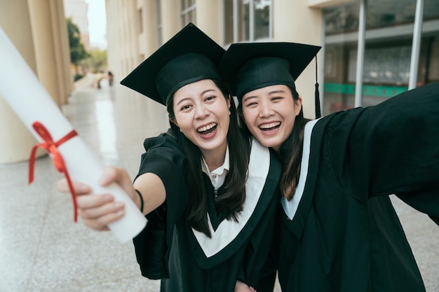 podekscytowani dwaj szczęśliwi absolwenci, którzy odnieśli sukces w szatach i czapkach z frędzlami, robiąc sobie zdjęcie stojące w sali budynku uniwersyteckiego. młode dziewczęta uczennice pokazujące dyplom ukończenia studiów blisko wziąć selfie