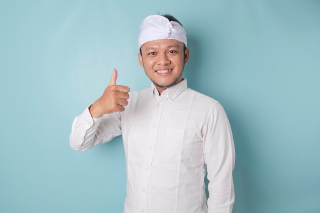 Podekscytowany Balijczyk noszący udeng lub tradycyjną opaskę na głowę i białą koszulę pokazuje kciuk w górze gest aprobaty na białym tle na niebieskim tle