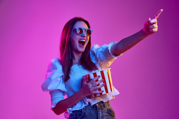 Podekscytowany film Portret młodej emocjonalnej dziewczyny pozuje w okularach 3D z koszem popcornu na różowym tle w neonowym świetle Koncepcja emocji młodzieżowego stylu życia