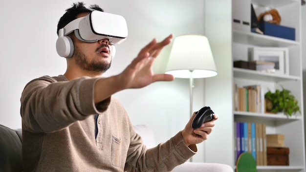 Podekscytowany i zdumiony. Młody Azjata noszący gogle VR podczas grania w gry wideo z rękami wyciągniętymi, by dotknąć czegoś w wirtualnym świecie.