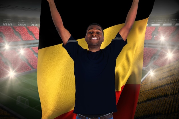 Podekscytowany kibic piłki nożnej w czarnym dopingu trzymający flagę belgii przed rozległym stadionem piłkarskim z fanami w kolorze żółtym i czerwonym