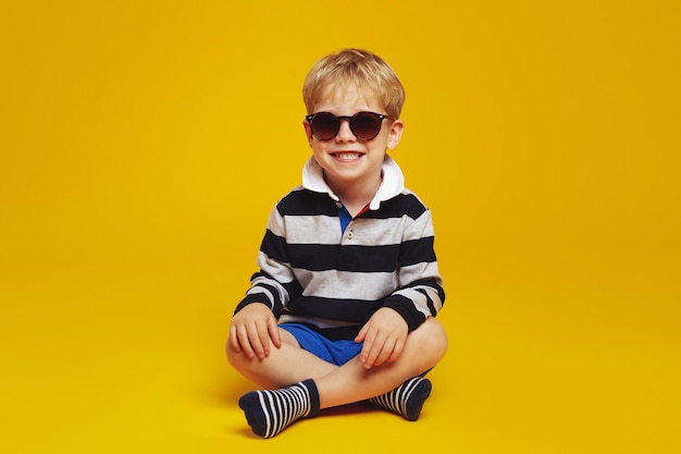 Podekscytowany mały chłopiec noszący modne okulary przeciwsłoneczne, siedząc ze skrzyżowanymi nogami na podłodze studia