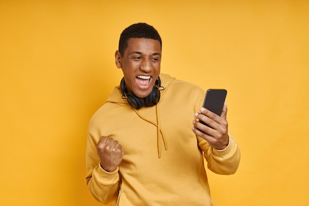 Zdjęcie podekscytowany mężczyzna rasy mieszanej ze słuchawkami, używający smartfona, stojąc na tle żółtego