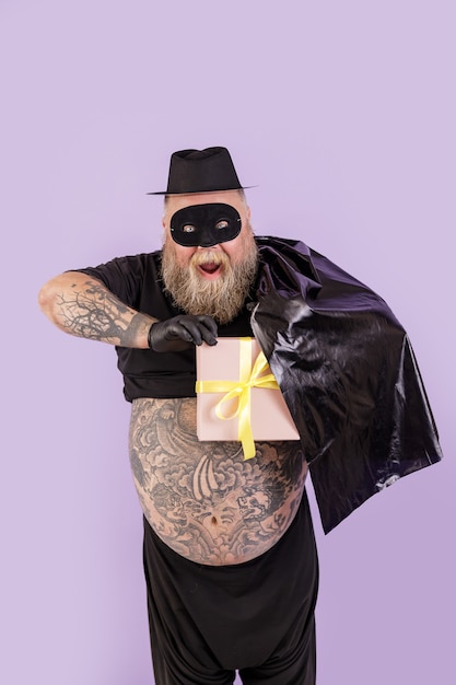 Podekscytowany mężczyzna z nadwagą w kostiumie zorro chowa pudełko za peleryną na fioletowym tle