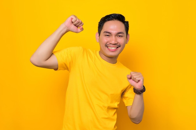Podekscytowany młody azjatycki człowiek w casual tshirt świętuje wielkie szczęście i sukces z uniesionymi pięściami na białym tle na żółtym tle Koncepcja życia ludzi