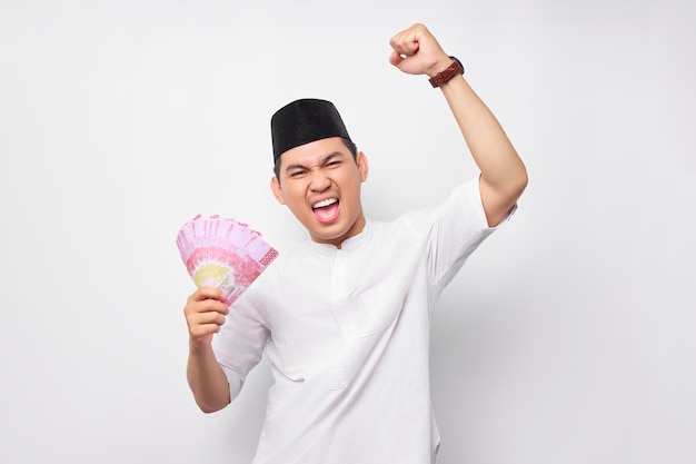 Podekscytowany młody azjatycki muzułmanin trzymający banknoty rupii w gotówce i świętujący sukces na białym tle Koncepcja stylu życia ludzi religijnych islamu