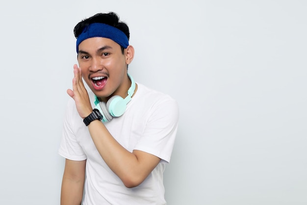 Podekscytowany młody azjatycki sportowiec w niebieskim pałąku i białej koszulce sportowej ze słuchawkami szepczącymi plotki i dzielącymi się wiadomościami na białym tle Koncepcja sportowa treningu