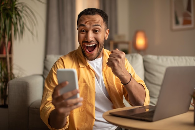 Zdjęcie podekscytowany młody czarny mężczyzna patrzący na ekran smartfona, gestykulujący tak, zadowolony z wygranej online w