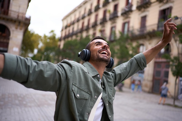 Podekscytowany młody człowiek z słuchawkami stoi na europejskiej ulicy Barcelony z wyciągniętymi ramionami