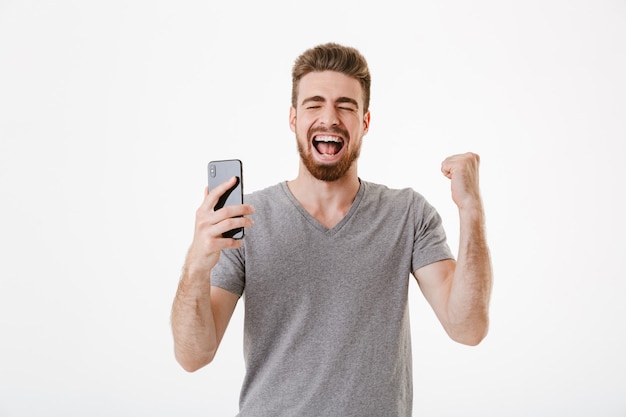 Podekscytowany młody człowiek za pomocą telefonu komórkowego robi gest zwycięzcy.