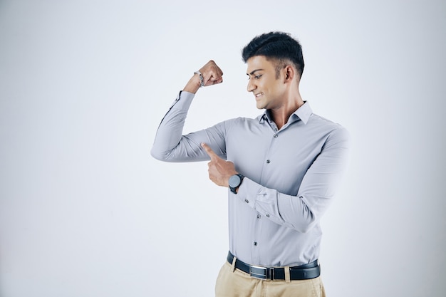 Podekscytowany młody indyjski biznesmen wskazujący na jego twarde mięśnie bicepsa, na białym tle