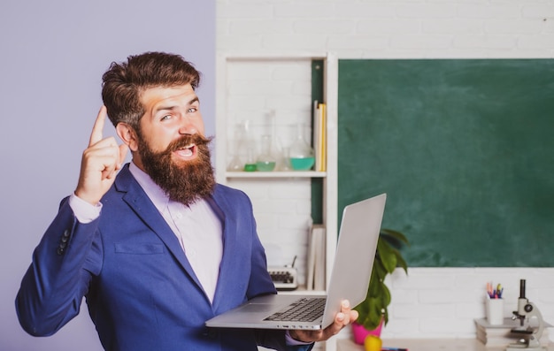 Podekscytowany nauczyciel z palcem wskazującym trzyma notebook laptopa w klasie w szkole zdumiony nauczyciel czeka