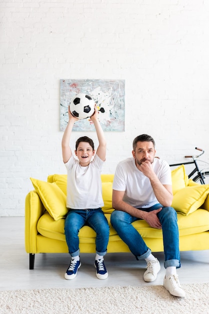 Podekscytowany syn z piłką nożną ogląda mecz sportowy z ojcem w salonie