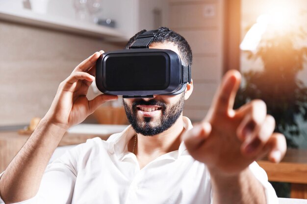 Podekscytowany wielokulturowy brodaty mężczyzna dopasowujący gogle wirtualnej rzeczywistości na głowie w nowoczesnym mieszkaniu