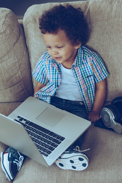 Podoba mi się ten laptop. Wysoki kąt widzenia małego afrykańskiego chłopca patrzącego na laptopa siedząc na kanapie w domu