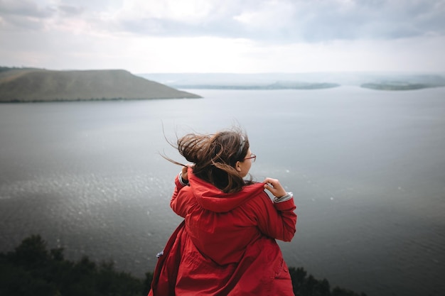 Podróżniczka w czerwonym płaszczu przeciwdeszczowym i wietrznych włosach stojąca na szczycie skalistej góry z pięknym widokiem na rzekę Młoda hipsterka relaksująca się na klifie Podróże i zamiłowanie do wędrówki Kopiuj przestrzeń