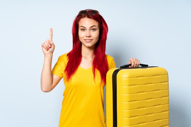 Podróżnik dziewczyna trzyma walizkę odizolowywająca na błękit ścianie wskazuje w górę doskonałego pomysłu