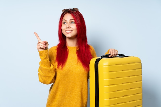 Podróżnik kobieta trzyma walizkę odizolowywająca na błękit ścianie wskazuje w górę doskonałego pomysłu
