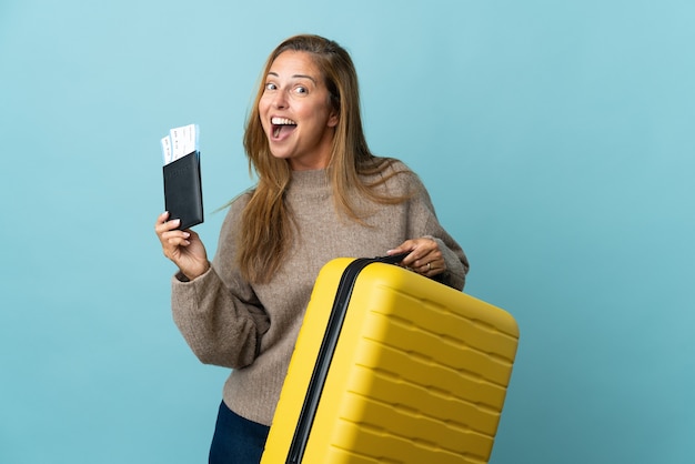 Podróżnik kobieta w średnim wieku trzyma walizkę na białym tle na niebieskiej ścianie w wakacje z walizką i paszportem