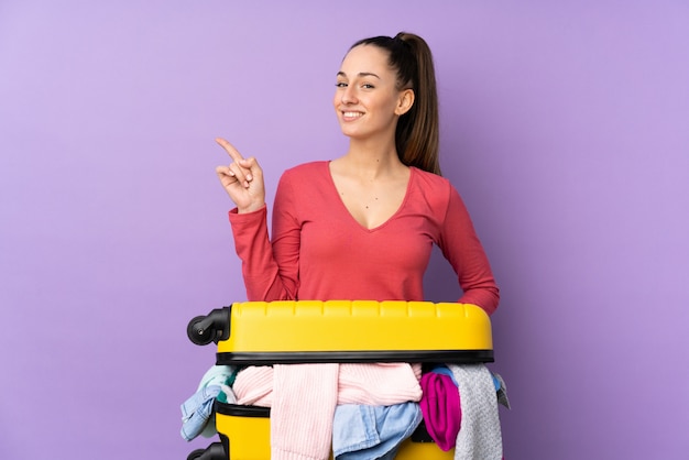 Podróżnik kobieta z walizką pełną ubrań na pojedyncze fioletowe ściany palcem wskazującym na bok