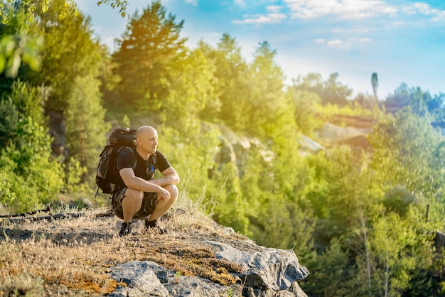 Podróżnik patrzy na otaczający go krajobraz na szczycie klifu latem przy ciepłej pogodzie Mężczyzna siedzi na przysiadzie przy klifie w słoneczny dzień