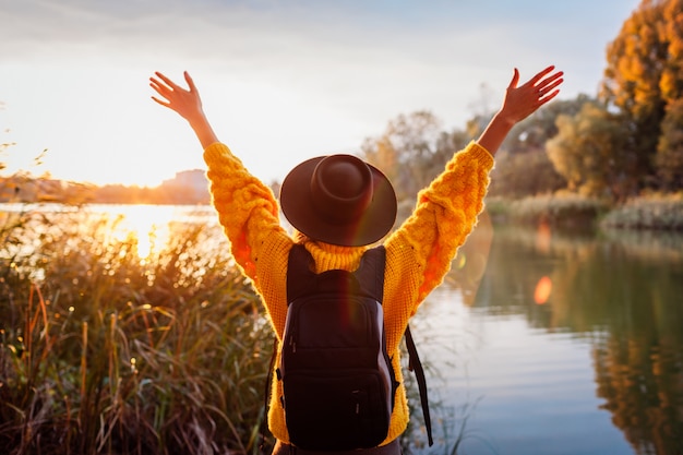 Zdjęcie podróżnik relaksuje jesień rzeką przy zmierzchem z plecakiem. młoda kobieta podniósł ręce czując się swobodnie i szczęśliwy