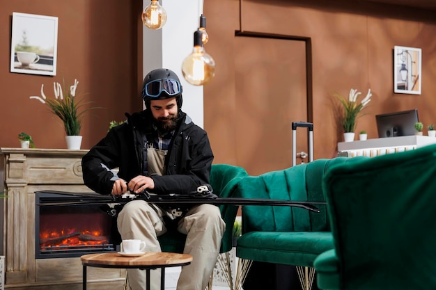 Podróżnik ubrany w kurtkę zimową dostosowuje sprzęt do sportów zimowych w hotelowym lobby, przygotowując się na śnieżną przygodę w ośrodku narciarskim. Kaukaski mężczyzna niosący i sprawdzający narty w salonie.