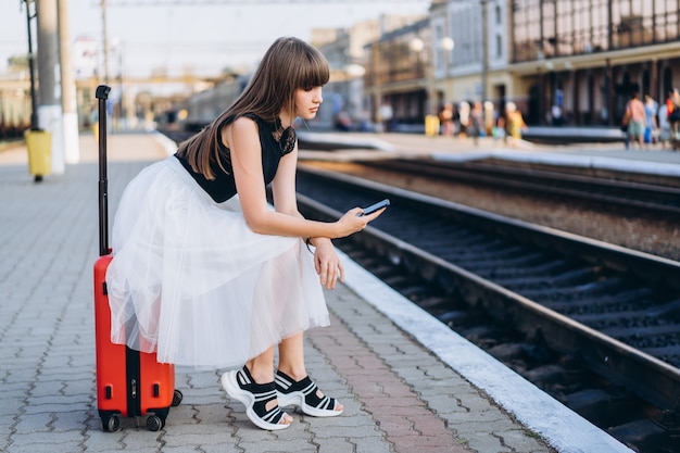 Podróżnik z czerwoną walizką w białej spódnicy czekający na pociąg na dworcu