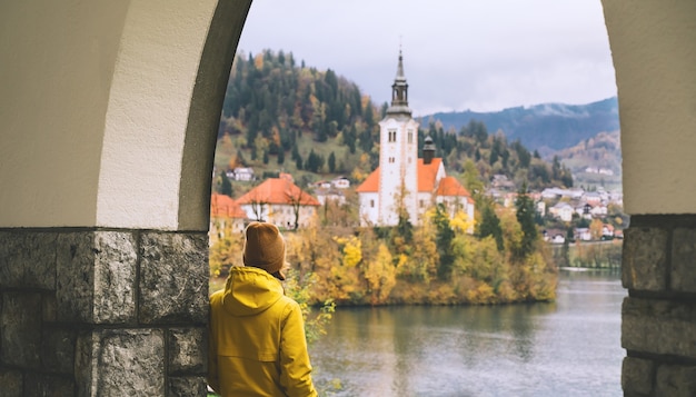 Podróżuj po Europie Turysta w żółtym płaszczu przeciwdeszczowym, patrząc na wyspę z kościołem na jeziorze Bled w Słowenii
