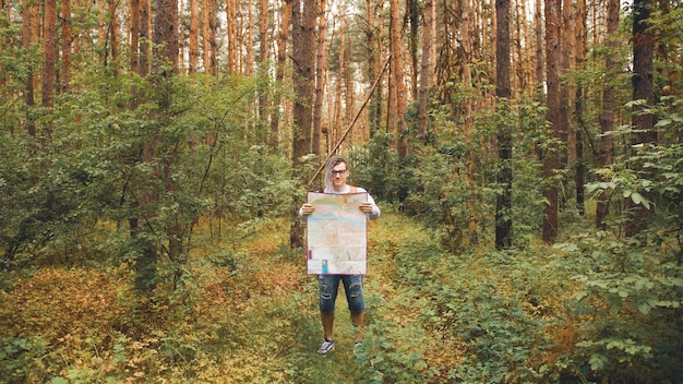 Podróżujący mężczyzna z dredami z mapą w lesie Mężczyzna podróżnik z plecakiem w lesie czyta mapę przy pochmurnej pogodzie