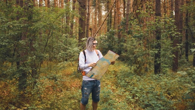 Podróżujący mężczyzna z dredami z mapą w lesie Mężczyzna podróżnik z plecakiem w lesie czyta mapę w pochmurną pogodę