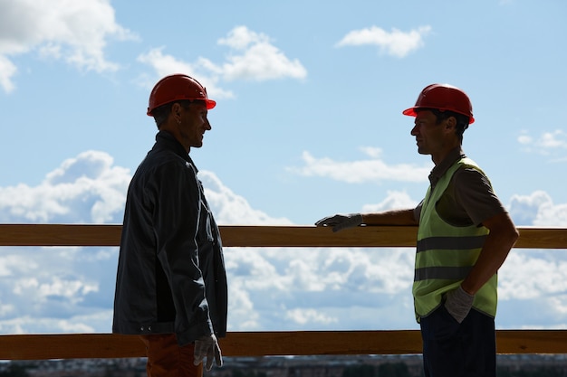 Podświetlany widok z boku dwóch pracowników budowlanych w kaskach i rozmawiających, stojąc na tle nieba, kopia przestrzeń