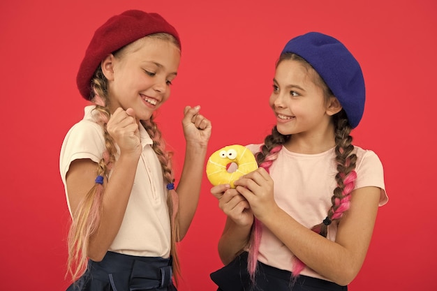Podziel się słodkim pączkiem Dziewczyny w beretach trzymają glazurowane pączki na czerwonym tle Dzieci figlarne dziewczyny gotowe zjeść pączka Przyjaźń i hojność Koncepcja sklepu ze słodyczami i piekarni Dzieci są wielkimi fanami pieczonych pączków