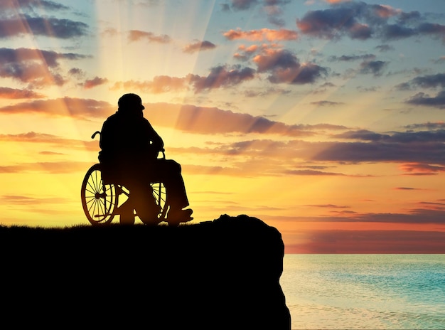 Pojęcie niepełnosprawności i starości. Sylwetka osoby niepełnosprawnej na wózku inwalidzkim na wzgórzu na tle morza o zachodzie słońca