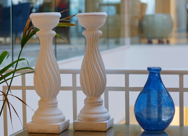 Pojedynczy niebieski wazon dwa białe świeczniki stoją na stole jako część dekoracji stołu Duże dekoracyjne świeczniki na stojaku na tle białej ściany i zielonej rośliny