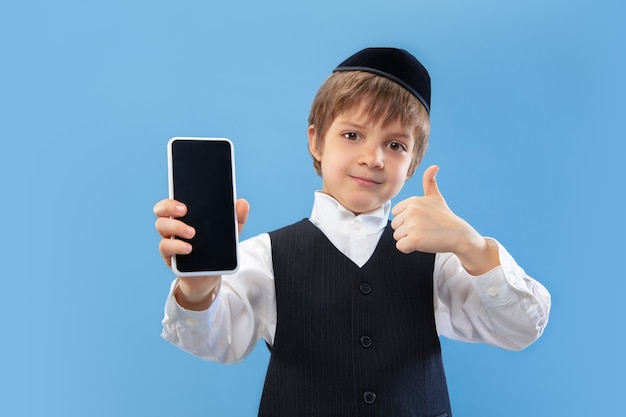 Pokazuje pusty ekran telefonu. Portret ortodoksyjnego żydowskiego chłopca na białym tle na ścianie niebieski studio.