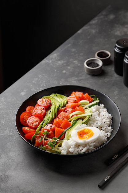 Poke bowl z łososiem, awokado, wiśnią, jajkiem i ryżem na ciemnym tle z pałeczkami.