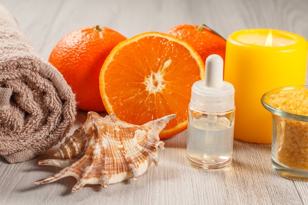 Pokrojona pomarańcza z dwoma całymi pomarańczami, ręcznikiem, muszelką, butelką z olejkiem do aromaterapii, szklaną miseczką z solą morską i palącą się świeczką na drewnianym biurku. Produkty i akcesoria spa.
